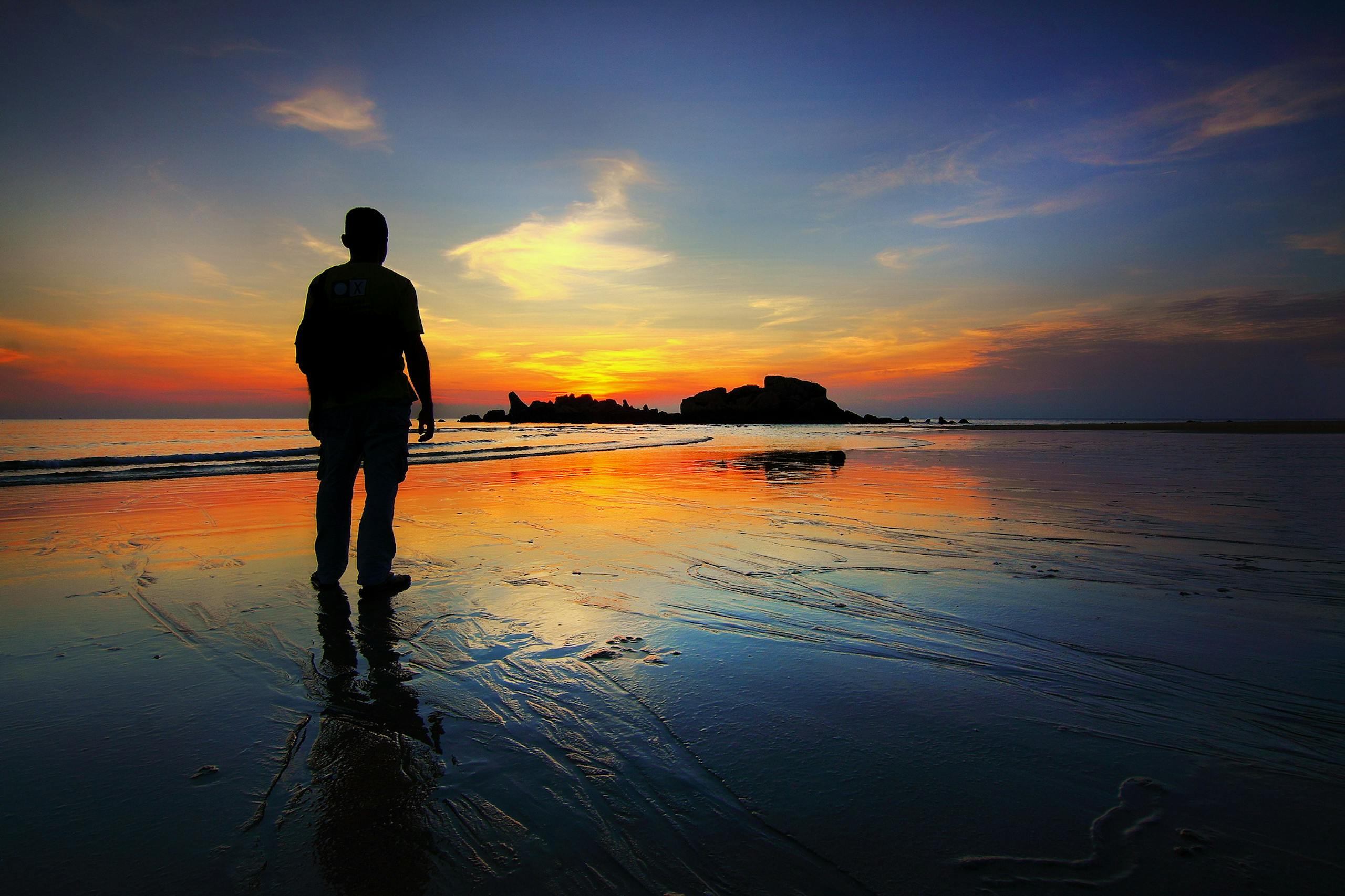 A silhouette of a person stands on the beach reflecting the vibrant sunset sky and tranquil sea. Finding Meaning over Metrics.
