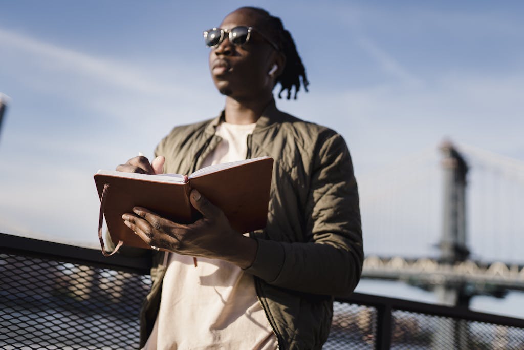 Man writing his personal story in a notebook near a bridge, enjoying a calm outdoor setting.